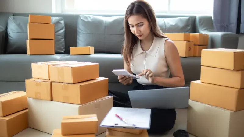 Mulher empreendedora mexendo no celular, com várias caixas de envio para e-commerce ao fundo. 